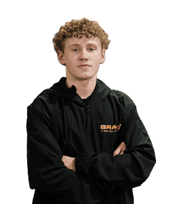 Young male athlete Noah Kelleher standing confidently with arms crossed, wearing a black jacket with "BRAY" logo, in an indoor sports facility.