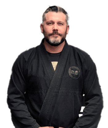 Home 8 - Bravé MMA And Fitness Male martial arts instructor with beard and slicked-back hair wearing a black gi, standing in front of a black and orange martial arts logo background.